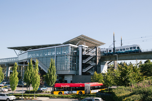 Link light rail leaving station via elevated tracks. A bus enters the station's ground-level parking lot.