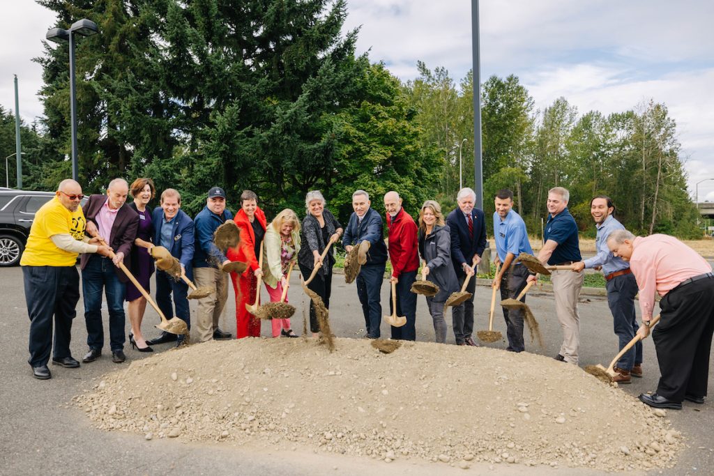 16 elected officials dig into a pile of dirt with shovels. Many of them are smiling.