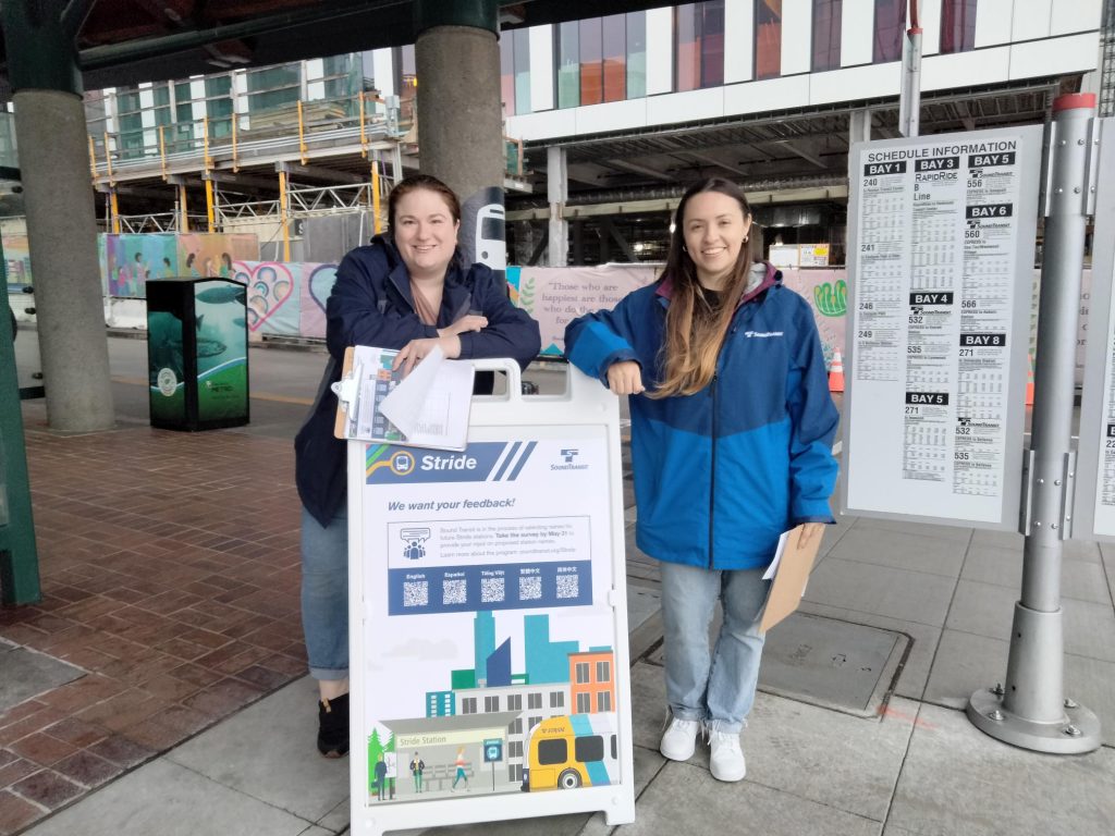 Two woman smile and stand behind an a-frame sign displaying Stride station naming information.